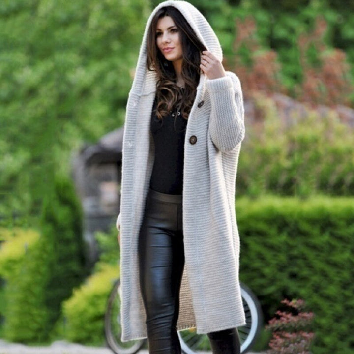 Eine Frau trägt eine lange beige Strickjacke mit Kapuze, dazu eine schwarze Innenhose und ein Oberteil