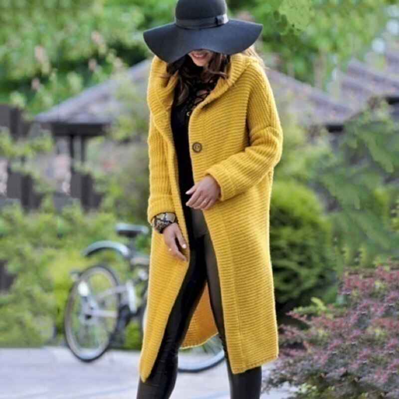 Eine Frau trägt eine lange gelbe Strickjacke mit Kapuze, dazu eine schwarze Innenhose und eine Mütze